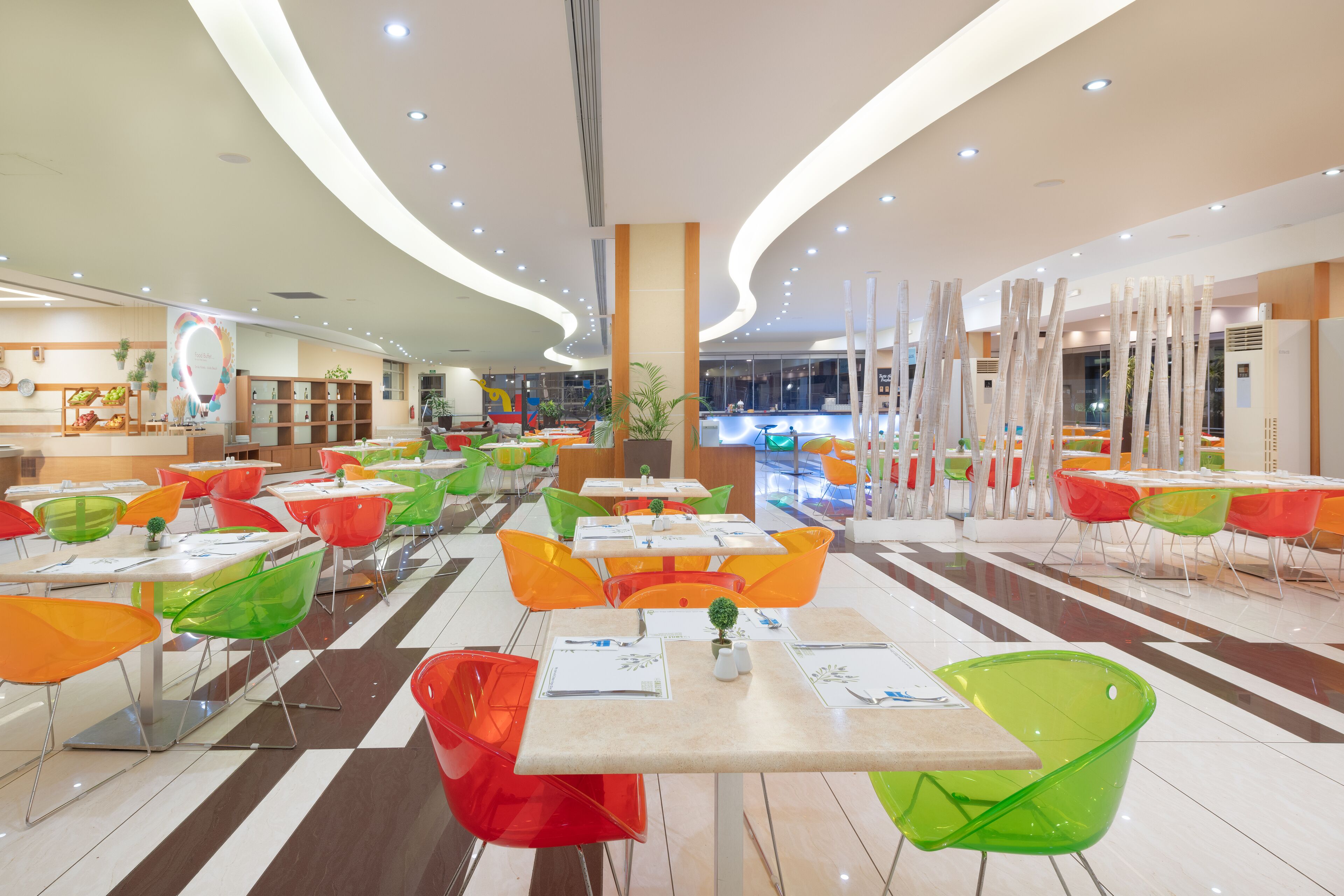 A modern dining area with colorful chairs and tables, a central plant, and a bright, well-lit ceiling.