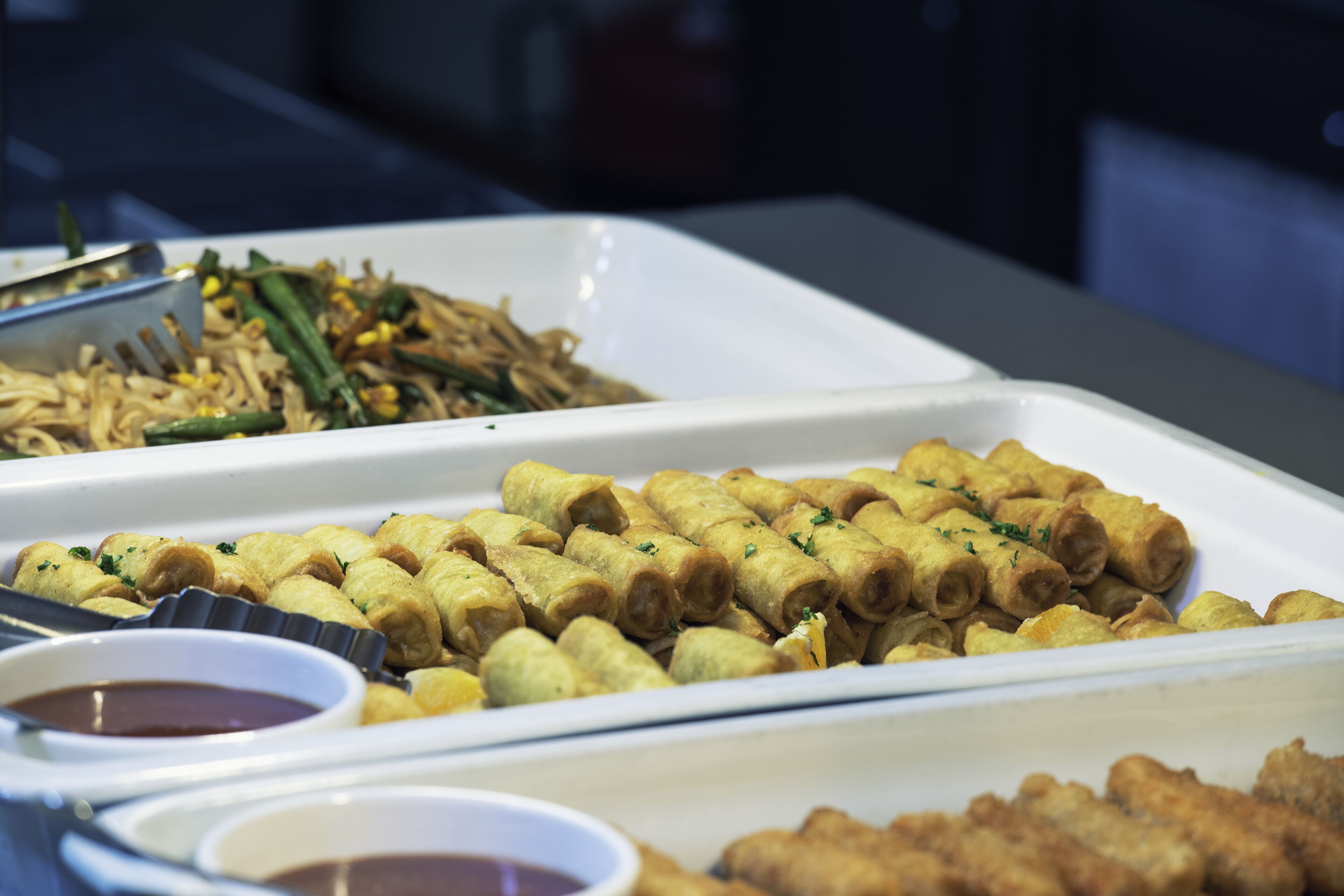 Un buffet con diversi piatti, tra cui involtini primavera, noodles e cibi fritti.