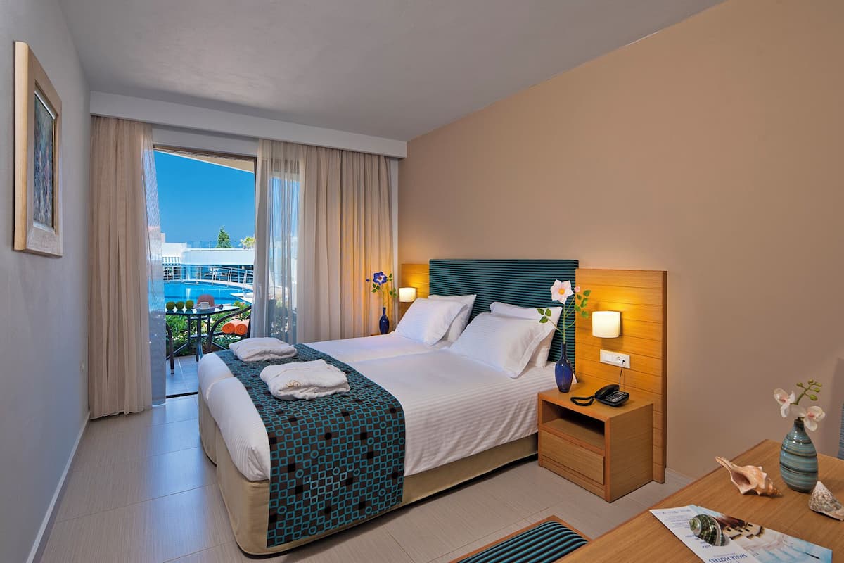 Habitación de hotel con cama, mesita de noche con lámpara, vista a la piscina y balcón.