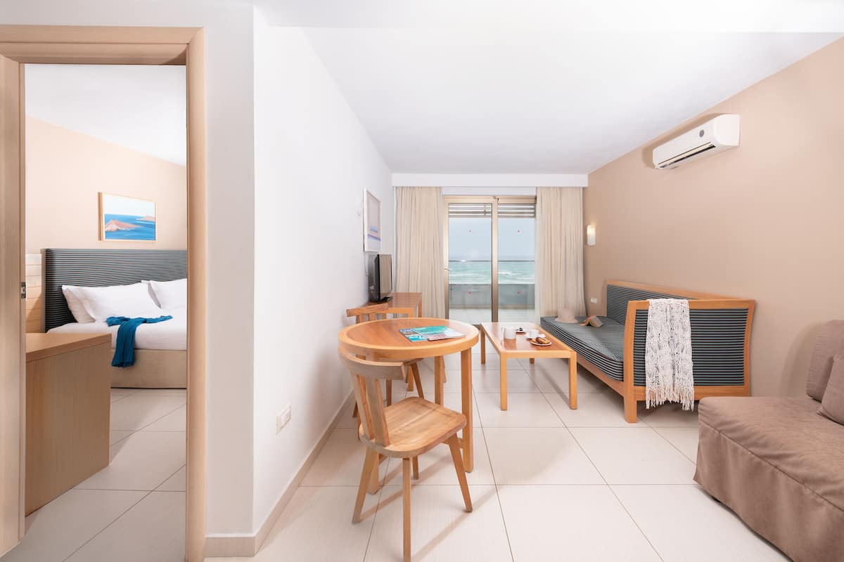 A modern hotel room with a bed, a sofa, a dining table with chairs, and a view of the sea through a large window.