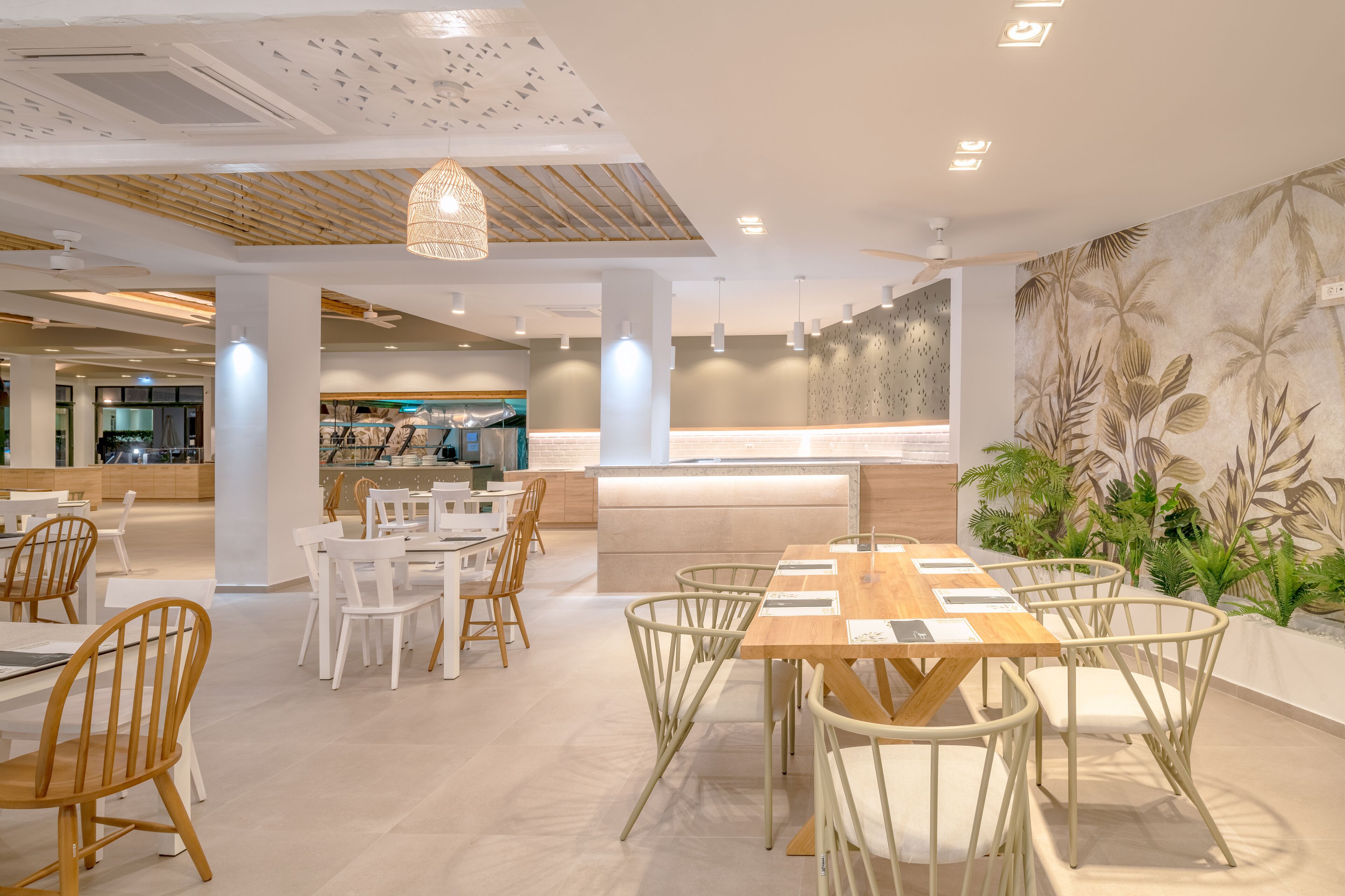 A modern dining area with wooden tables and chairs, a central island, and a wall with a tropical leaf pattern.
