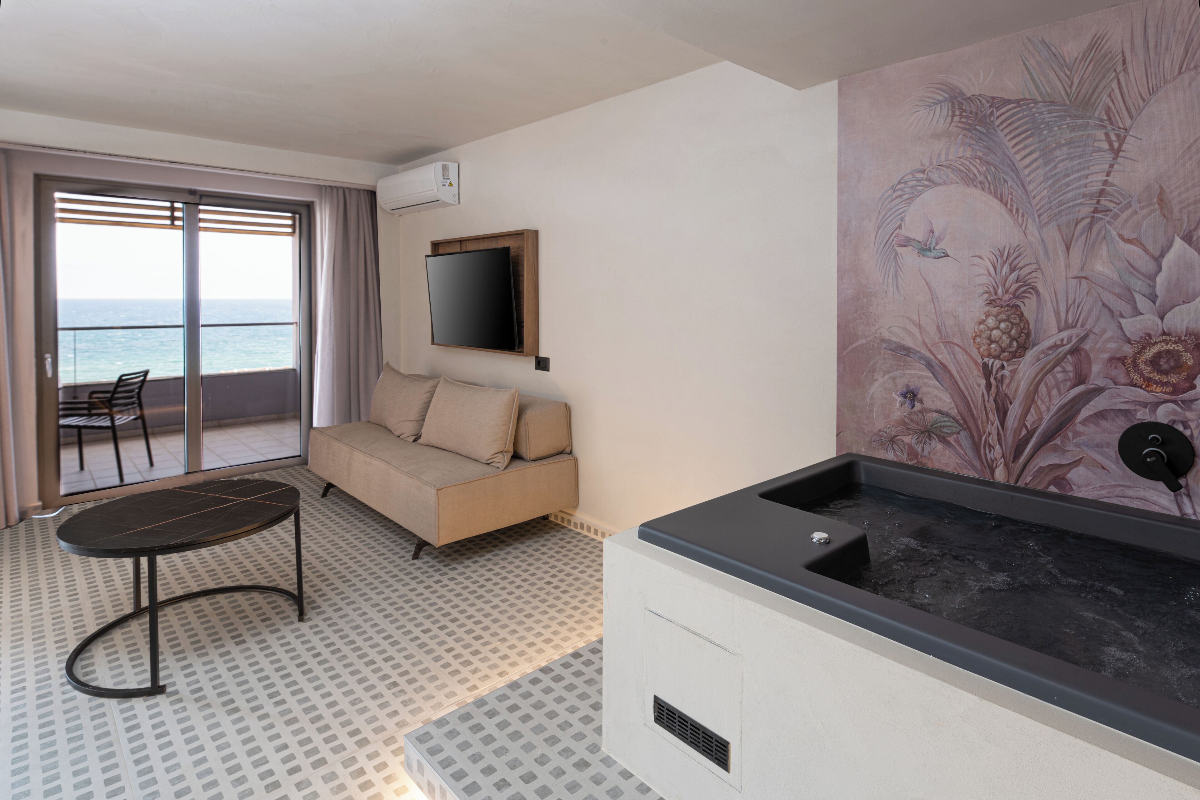 A modern room with a sofa, a small table, a large window with a view of the sea, a mounted flat-screen TV, and a jacuzzi.