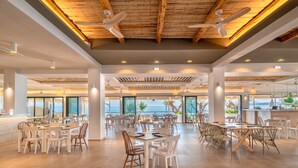 A spacious dining area with multiple tables and chairs, ceiling fans, and large windows offering a view of the sea. - Iolida Beach by Smile Hotels (Chania)