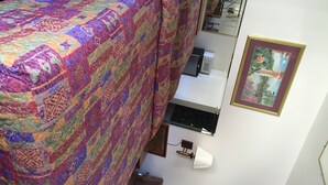 A bedroom with a bed covered in a colorful quilt, a small bedside table with a lamp, a mirror, and a framed picture on the wall. - Eagle Rock Motel (Los Angeles)