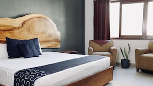 Un dormitorio con una cama grande, cabecera de madera, dos sillones, una planta en maceta y una ventana con cortinas.
