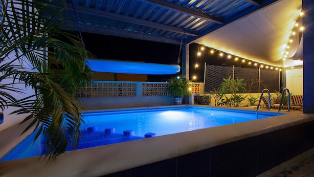 Una piscina con borde de baldosas, rodeada de plantas en macetas y una pérgola con luces colgantes.