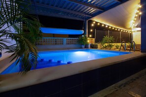 A swimming pool with a tiled deck, surrounded by potted plants and a pergola with string lights.