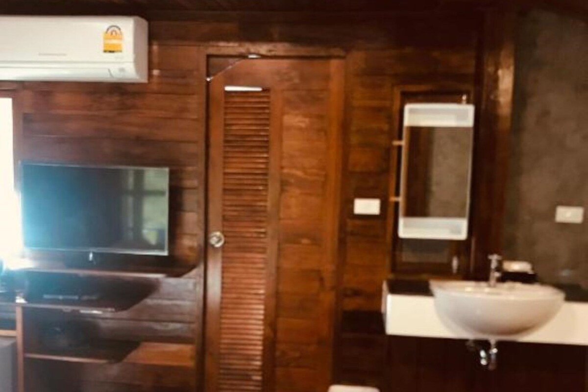 Une chambre d’hôtel avec un plancher en bois, un lit recouvert d’une couette blanche, un téléviseur fixé au mur, une porte en bois, un lavabo blanc et un miroir au-dessus du lavabo.