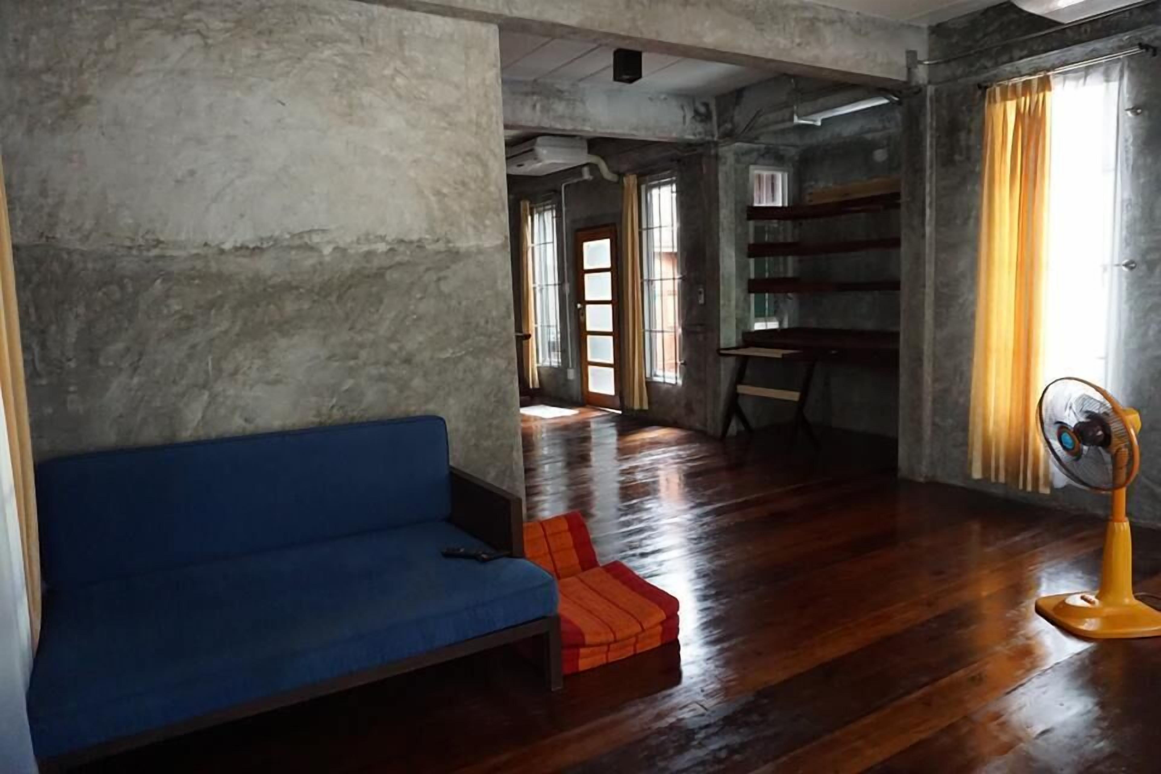 Une pièce avec un canapé bleu, un plancher en bois, un ventilateur jaune et une fenêtre avec des rideaux.