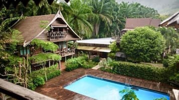 A traditional wooden house with a swimming pool surrounded by lush greenery.