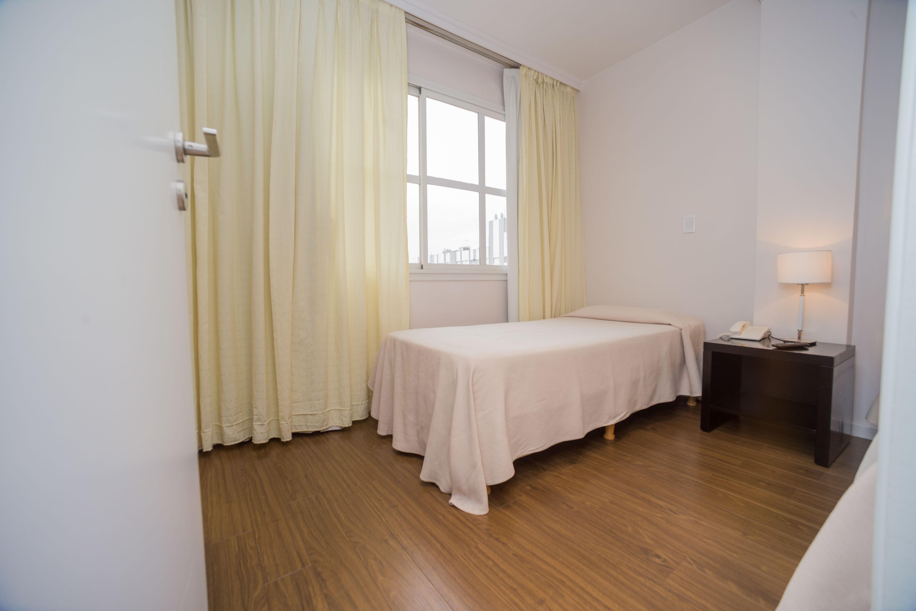 A room with a single bed, a nightstand with a lamp, and a window with curtains.