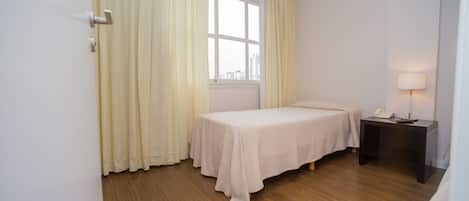 Una habitación con una cama individual, una mesita de noche con lámpara y una ventana con cortinas.