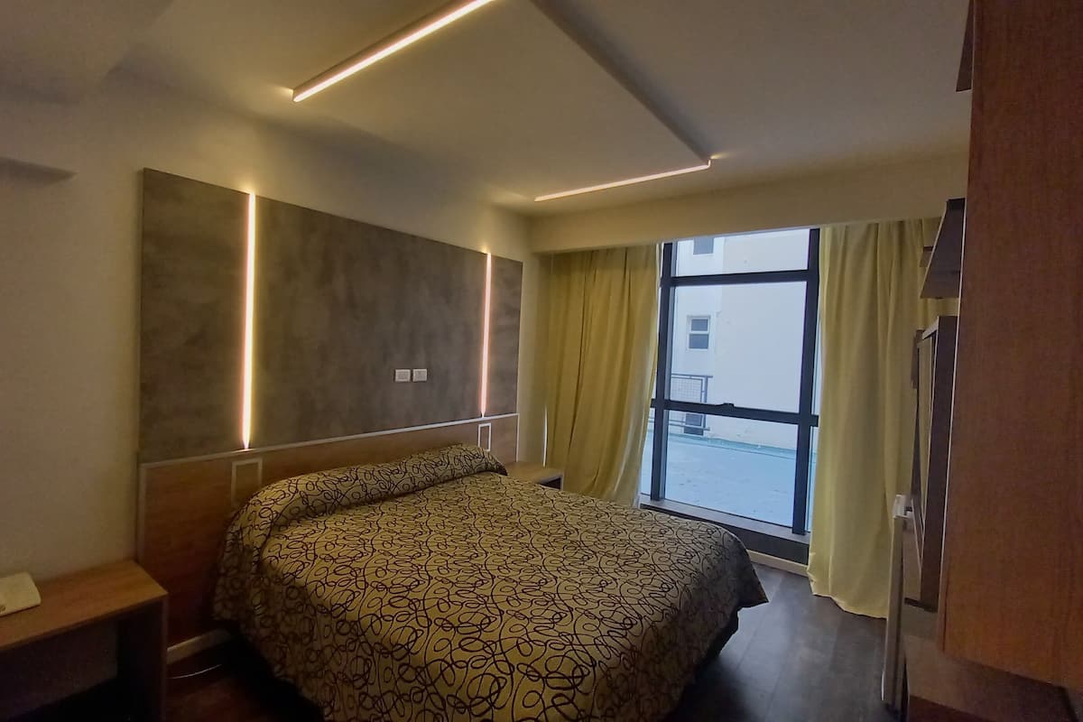 Una habitación de hotel moderna con una cama grande, una mesita de noche, un banco empotrado, una ventana con cortinas y vistas al exterior.