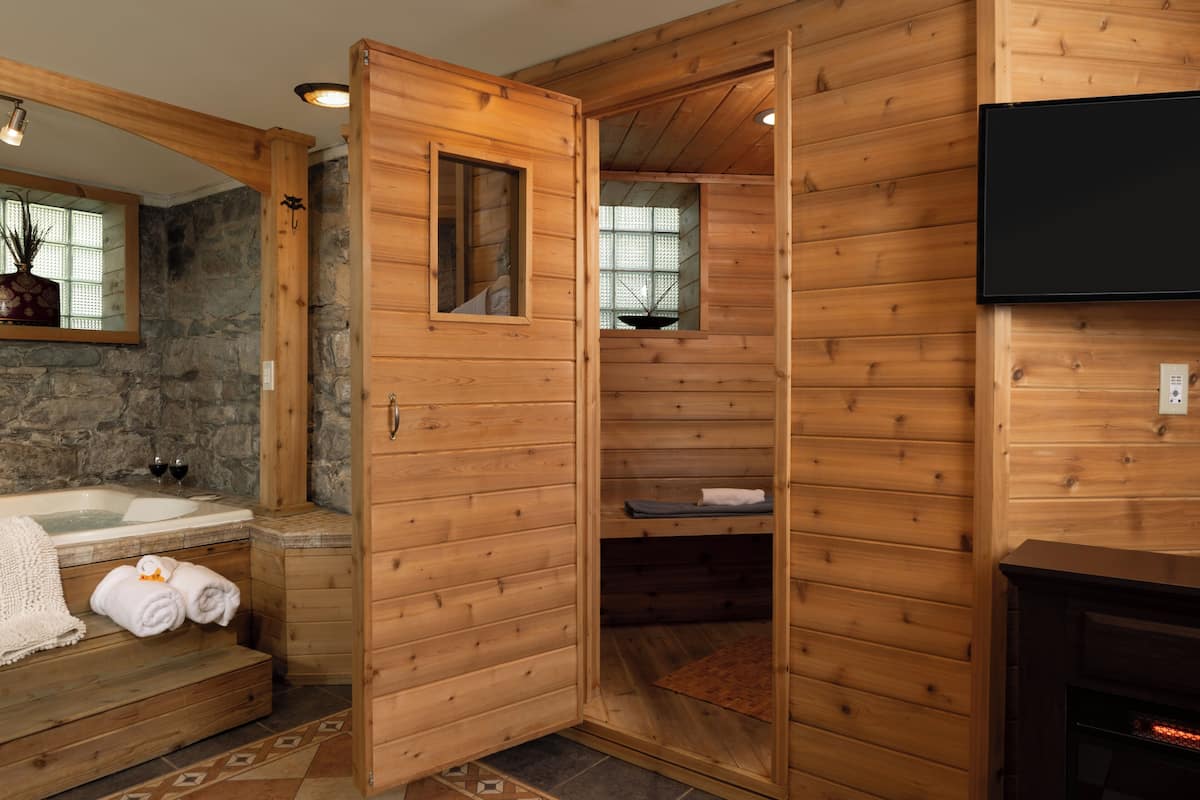 A wooden cabin interior with a stone wall, a wooden bathtub, a fireplace, and a television mounted on the wall.