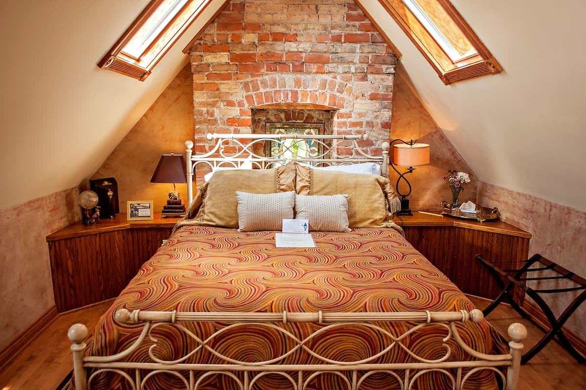 A bedroom with a brick fireplace, a bed with a patterned bedspread, two bedside tables with lamps, and a wooden nightstand.