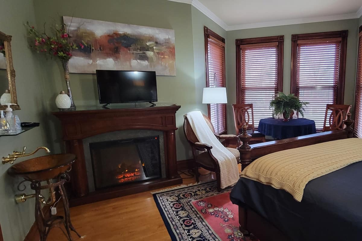 A bedroom with a fireplace, a television, a bed, a chair, and a window with blinds.