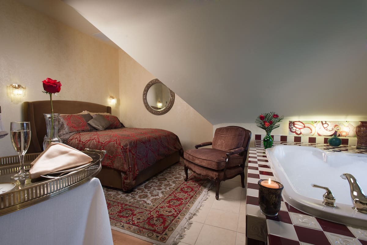 A bedroom with a bed, a chair, a bedside table with a glass, a vase with a rose, a mirror, a bathtub, and a checkered floor.