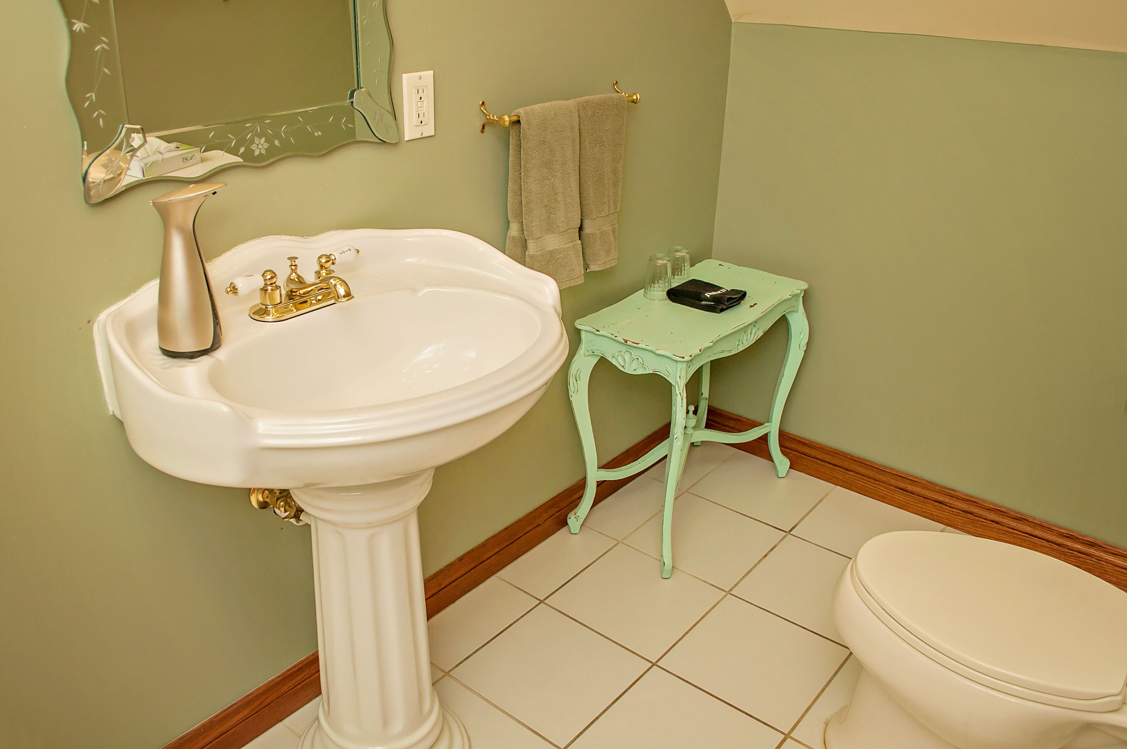 Un baño con lavamanos pedestal, un inodoro, una mesita con una toalla y un espejo en la pared.