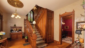 A grand staircase with ornate woodwork and a chandelier, leading to a dining area with a red wall and a sign for gourmet truffles. - The Historic Webster House (Bay City)
