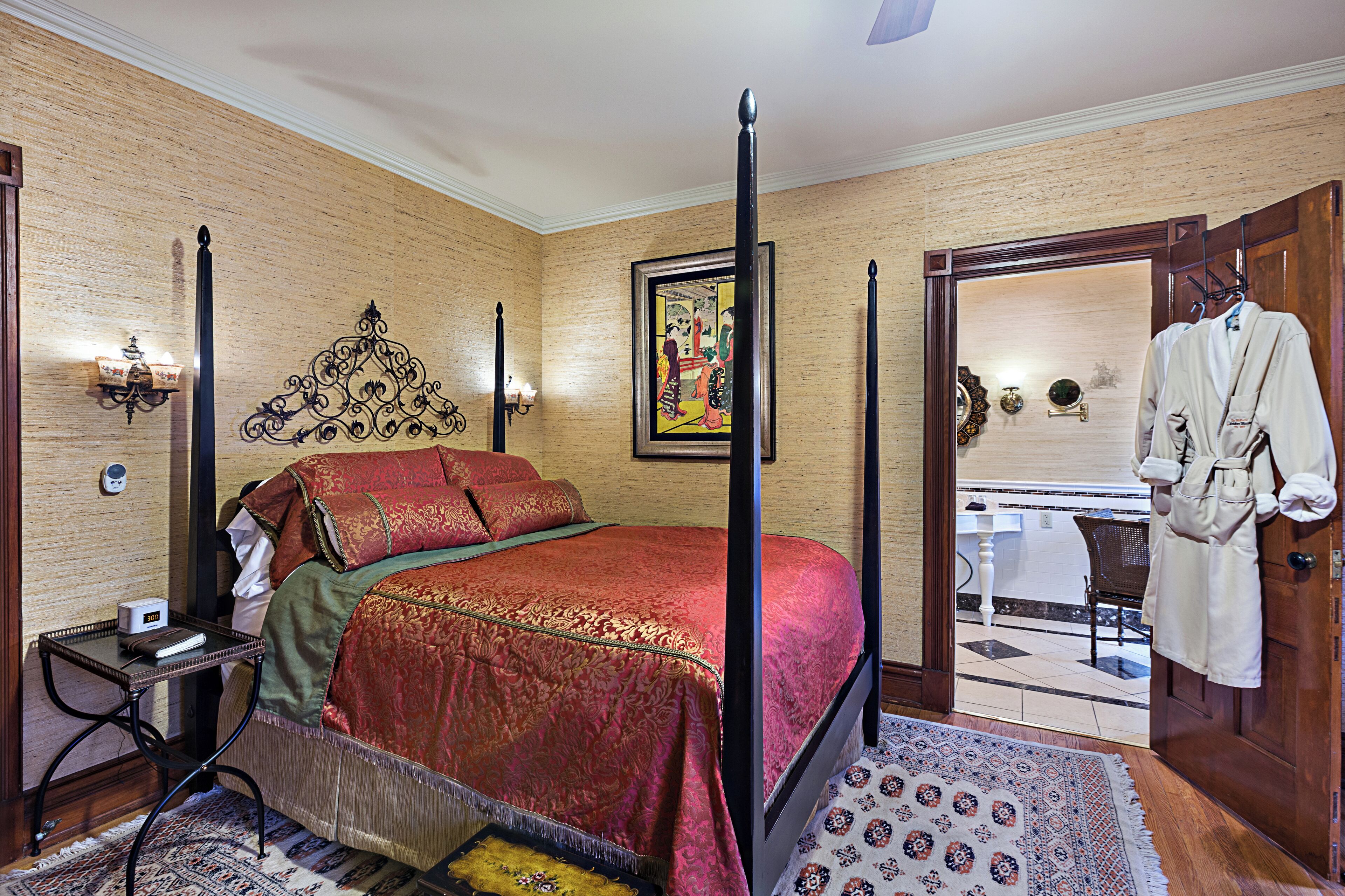 A four-poster bed with a red bedspread, a nightstand with a lamp, and a painting on the wall.