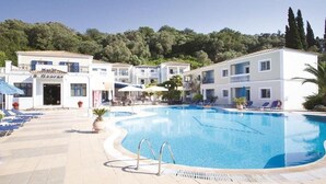 A swimming pool area with lounge chairs and umbrellas, surrounded by buildings with balconies and a view of a forested hillside. - San George (Corfu)