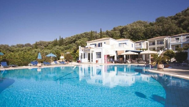 A clear blue swimming pool with lounge chairs and umbrellas, surrounded by buildings with a Mediterranean architectural style. - San George (Corfu)