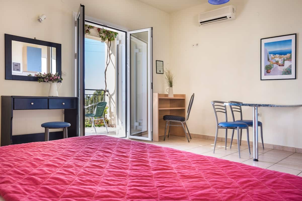 Un dormitorio con una colcha acolchada roja, un escritorio con dos sillas, un espejo y vistas al exterior a través de una puerta corrediza de vidrio.