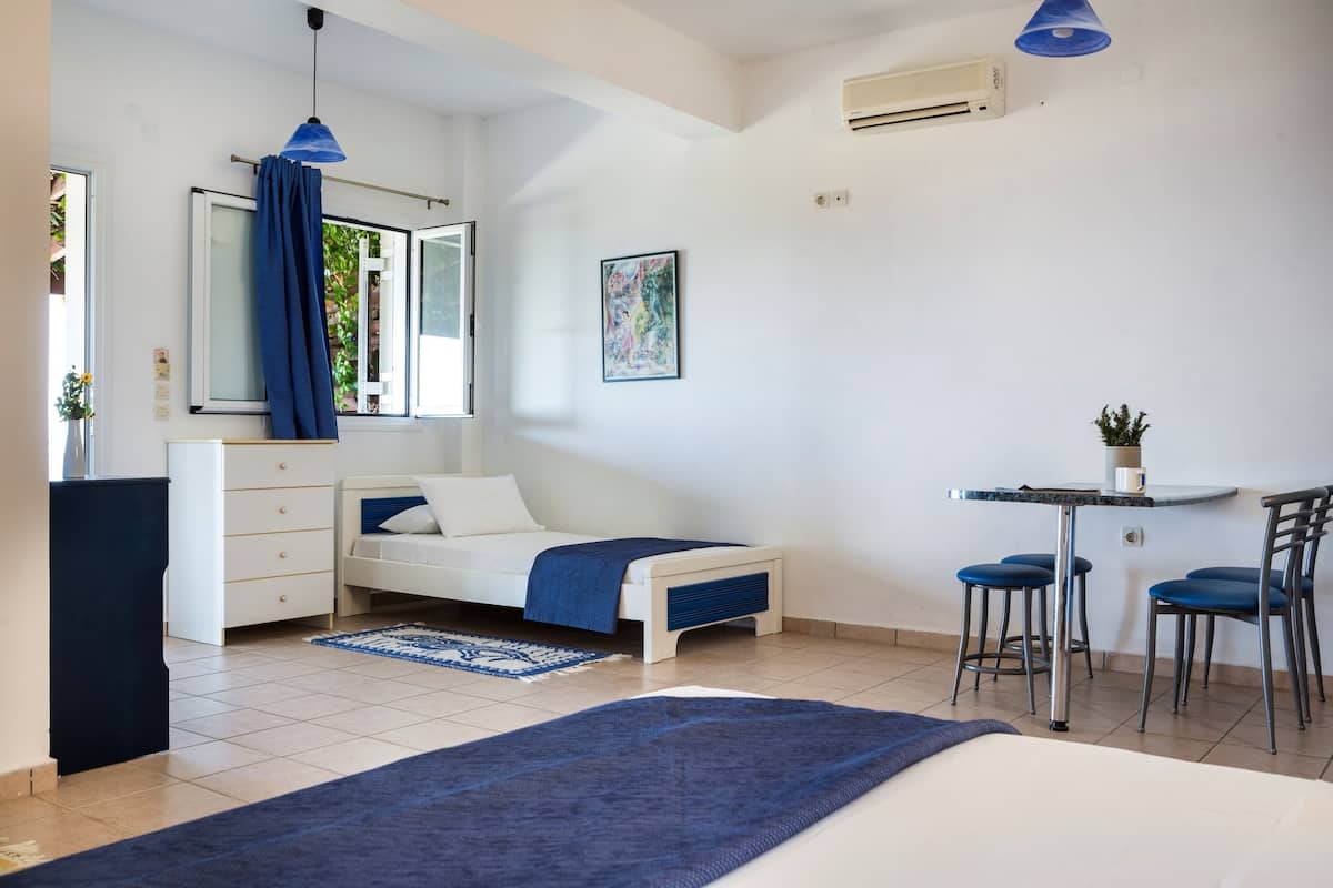 Un dormitorio con cama, escritorio, dos sillas, cómoda, ventana con cortinas y aire acondicionado.
