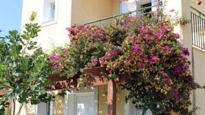 A two-story building with a balcony covered in flowering plants, a small patio with a table and chairs, and a door with a window.