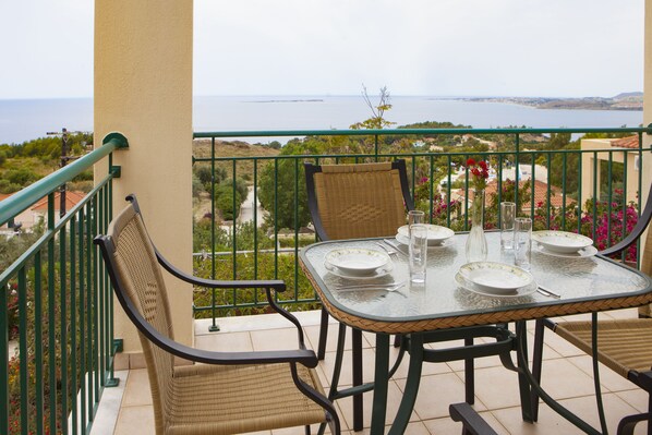 Un balcón con una mesa preparada para dos personas, con vista a un paisaje marino y vegetación.