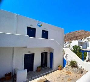 A white building with a blue sign, two windows, and a door. There are potted plants in front. - Hotel Naias (Serifos)