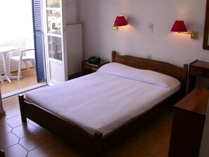 A hotel room with a bed, two bedside lamps, a bedside telephone, a wooden headboard, a nightstand, and a view of the outside patio. - Hotel Naias (Serifos)