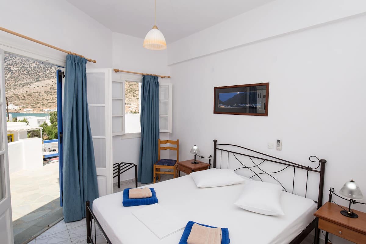 A bedroom with a bed, a chair, a nightstand, a lamp, and a view of the outside.