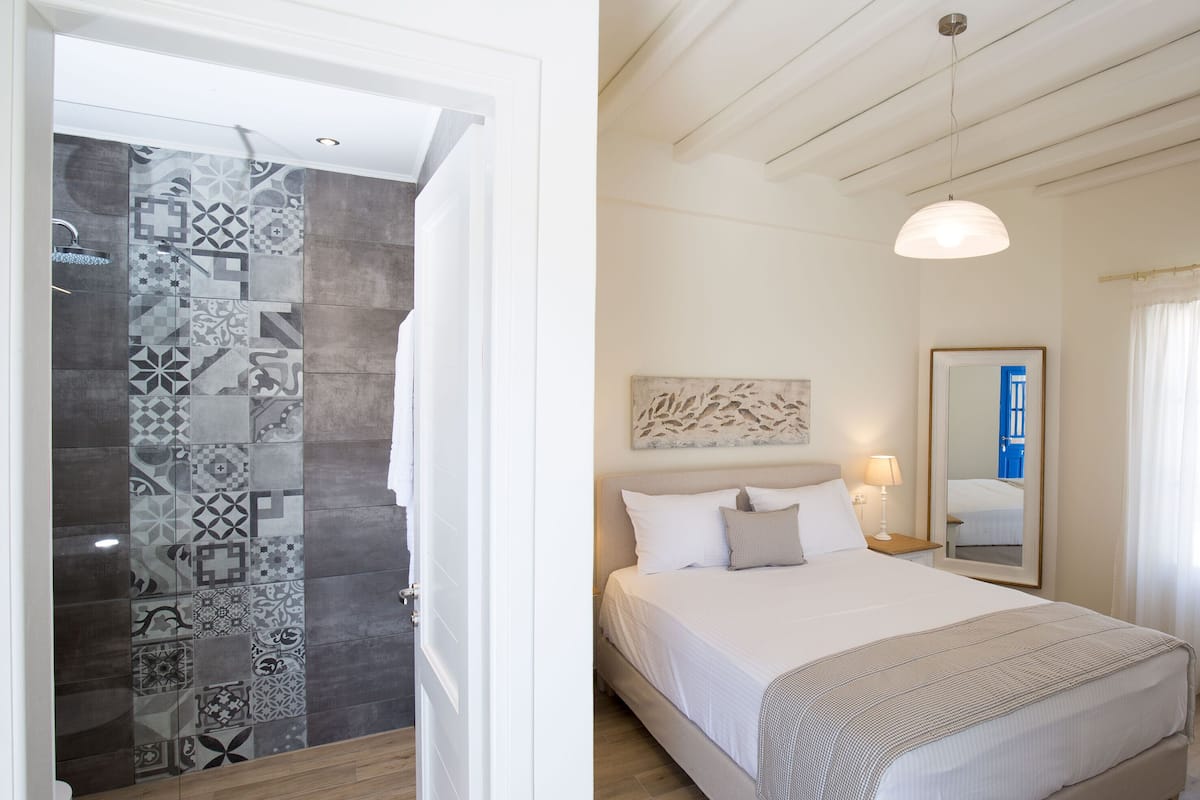 A bedroom with a bed, a bedside table, a lamp, a mirror, and a shower area with patterned tiles.