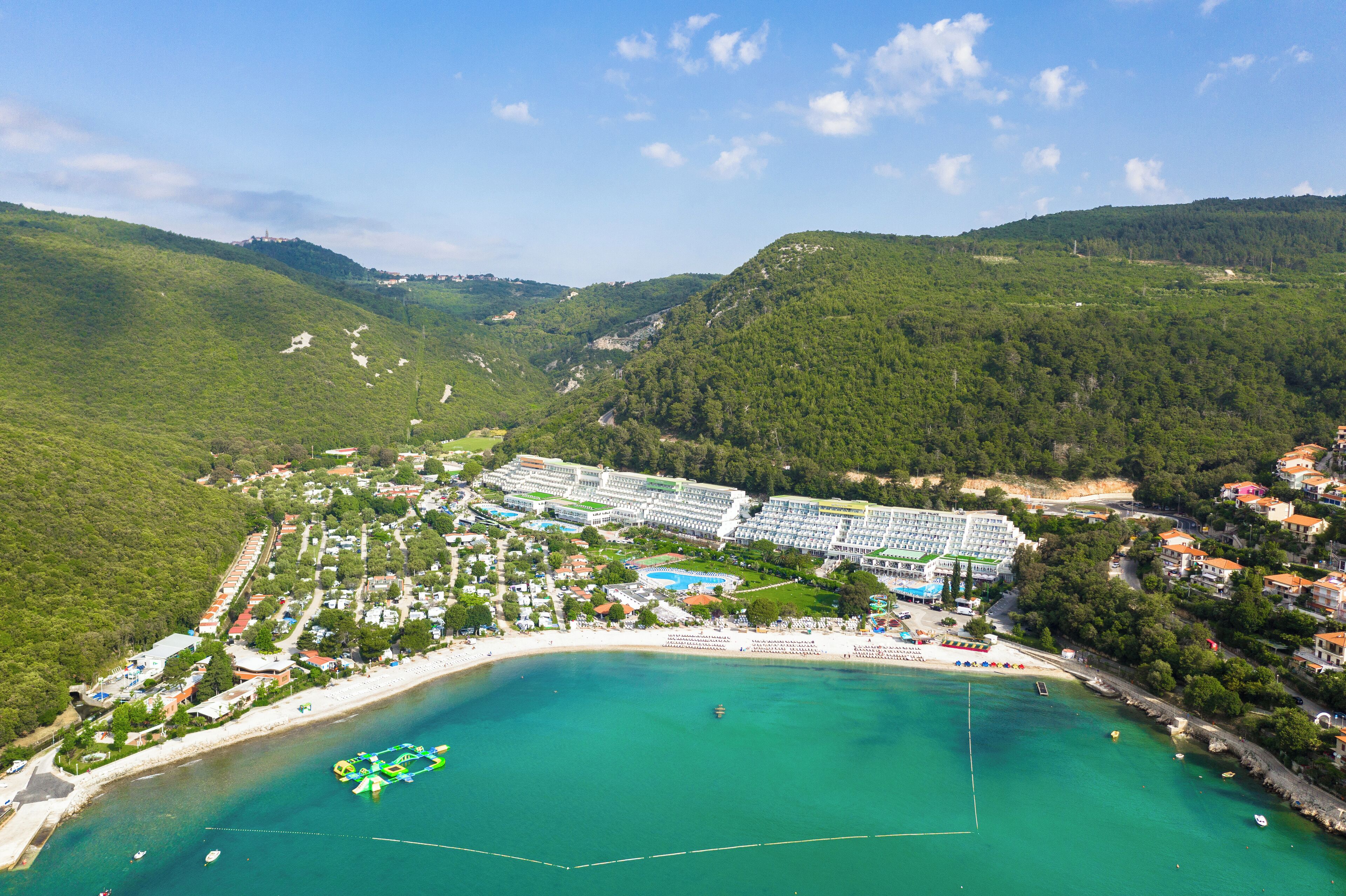 Luftaufnahme eines Küstenresorts mit mehreren Gebäuden, einem Strand und einer bergigen Kulisse.