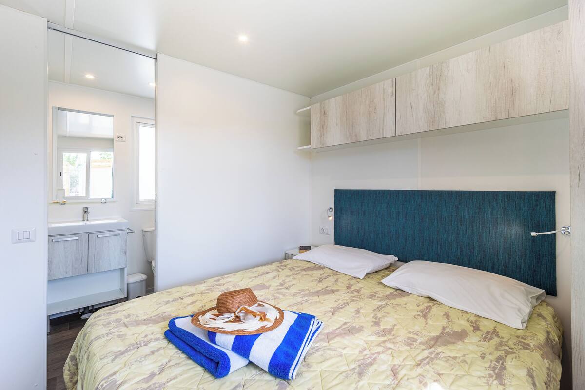 A bedroom with a bed, a headboard, a nightstand, a mirror, a bathroom with a sink and toilet, and a towel on the bed.