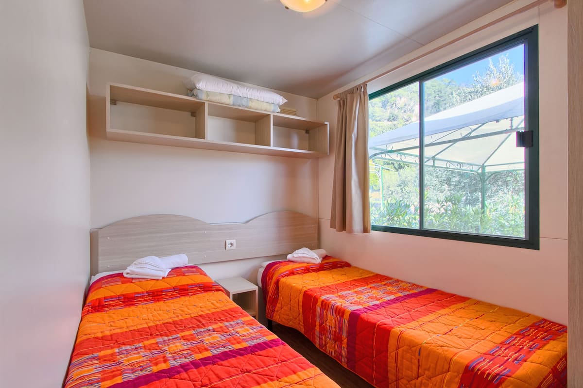 A room with two beds, a wooden headboard, a shelf above the beds, and a window with a view of greenery.