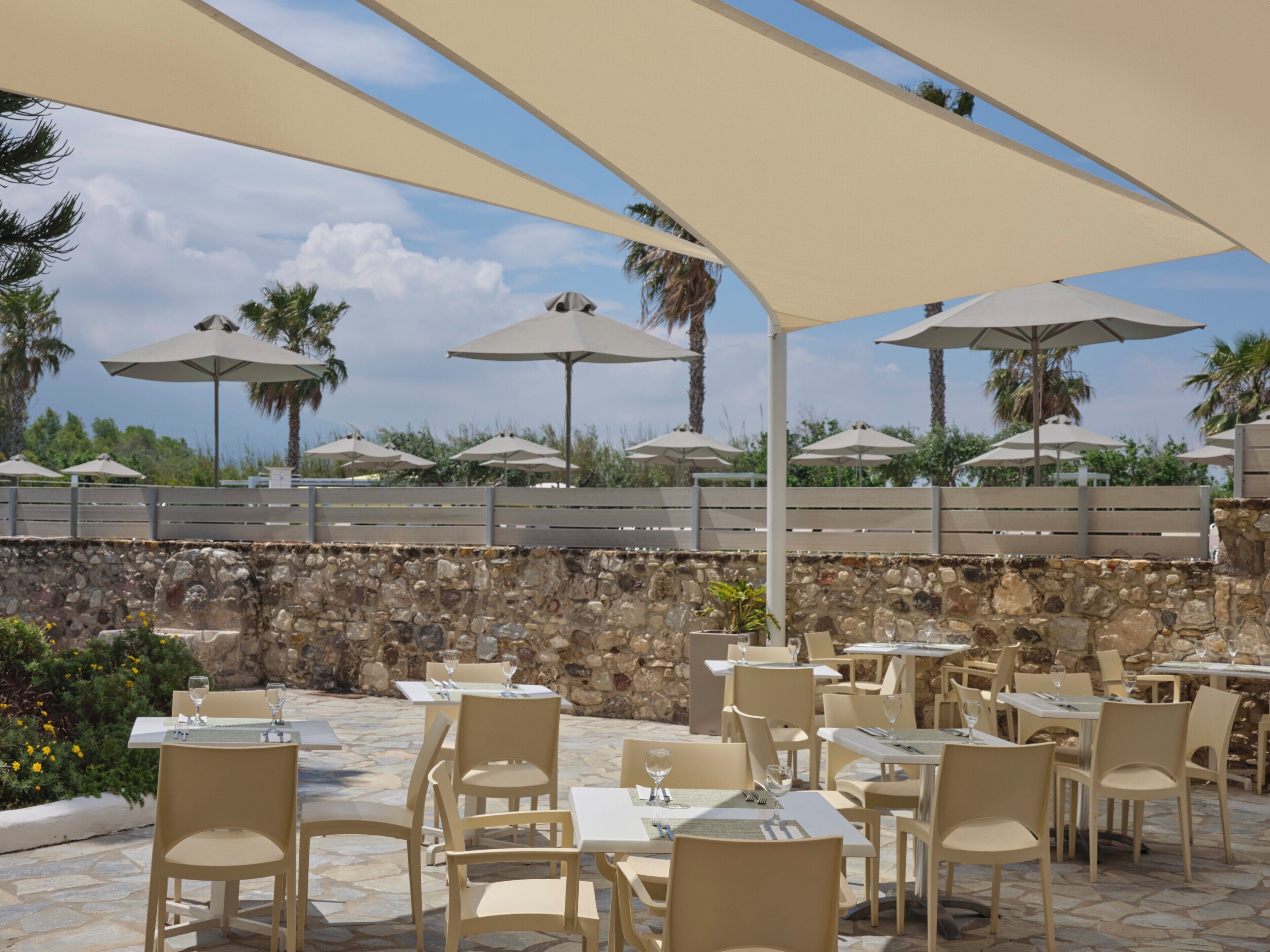 Un área de comedor al aire libre con mesas, sillas, sombrillas y una pared de piedra.