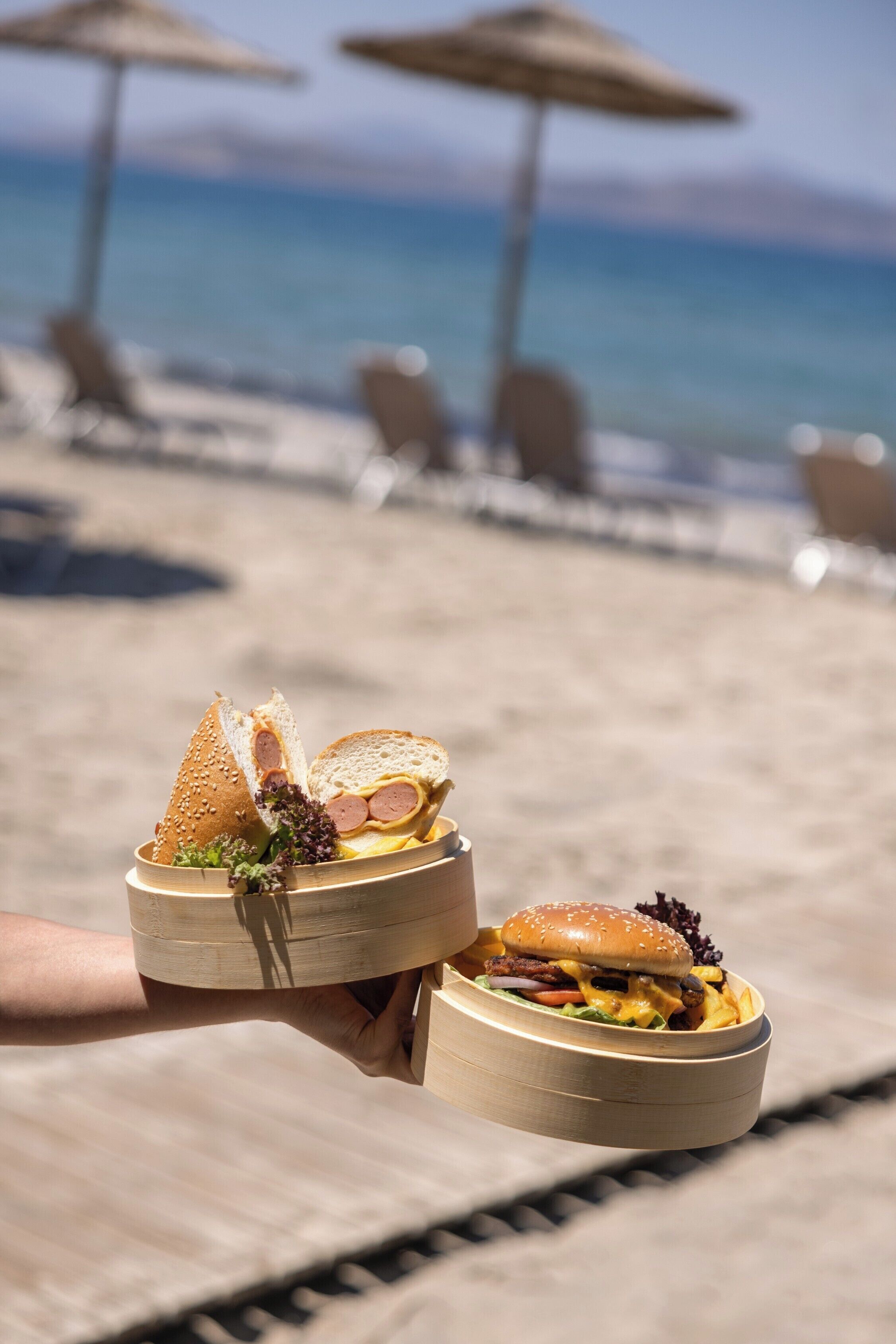 Una persona sosteniendo dos cuencos de madera con comida, un entorno de playa y sombrillas al fondo.