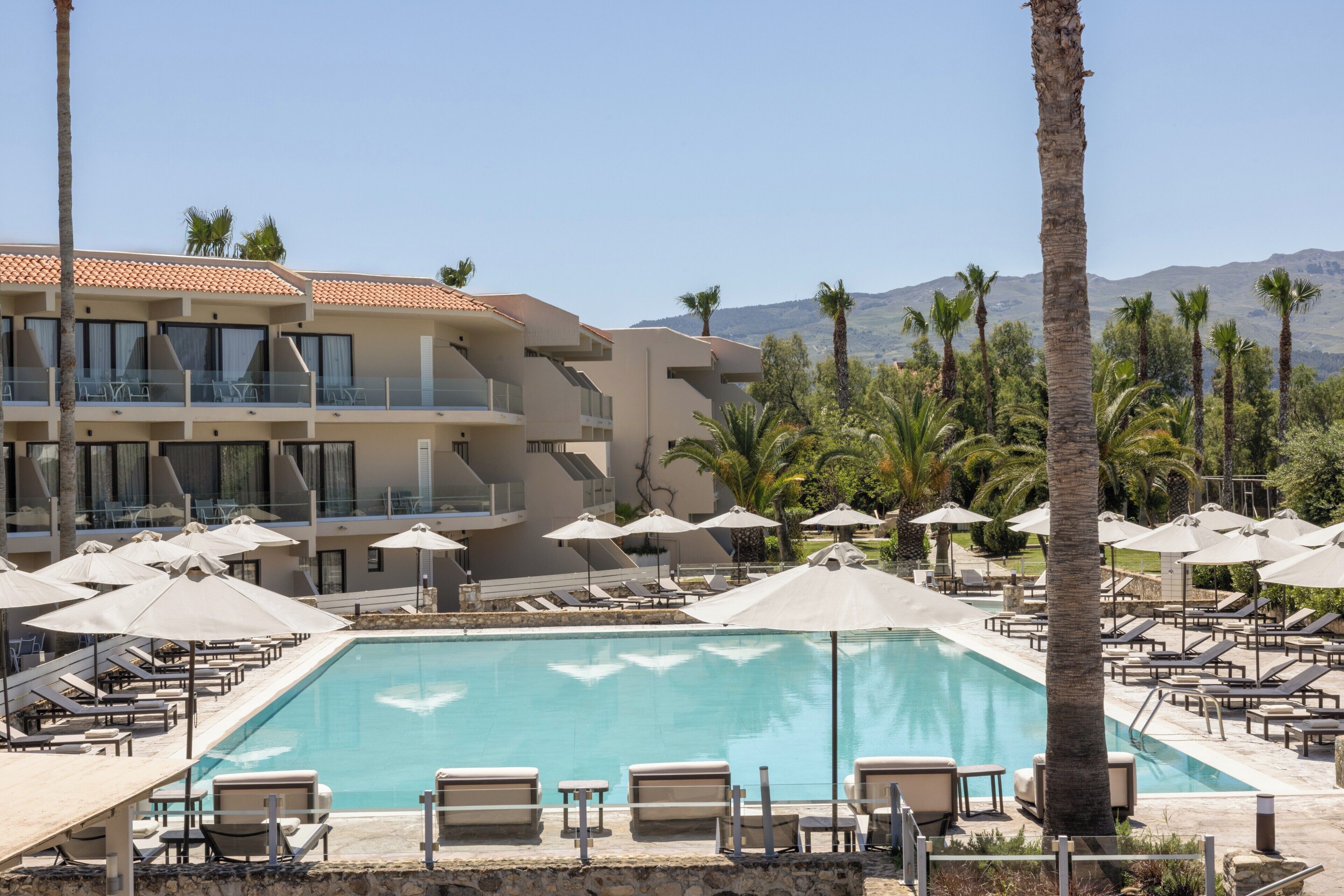 Un área de piscina con sillas de descanso y sombrillas, un edificio con balcones y palmeras.