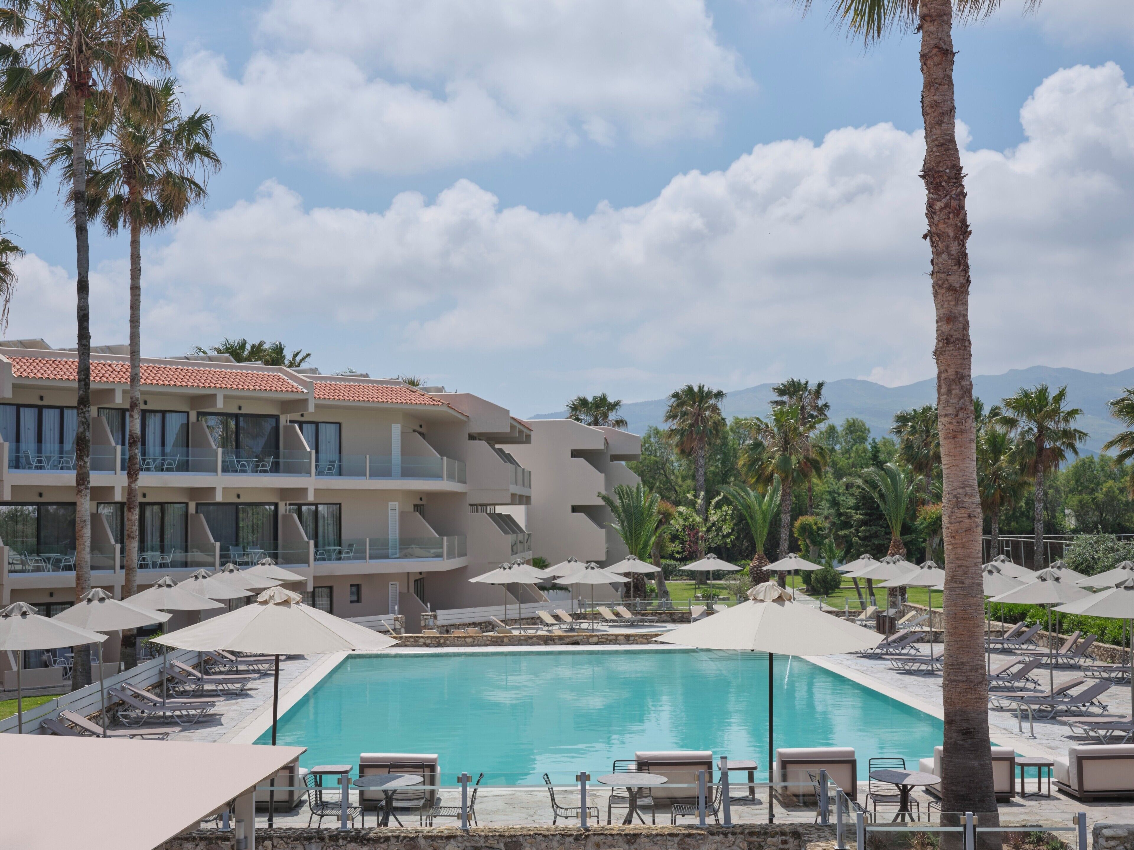 Un hotel con piscina, sillas de descanso, sombrillas y palmeras.