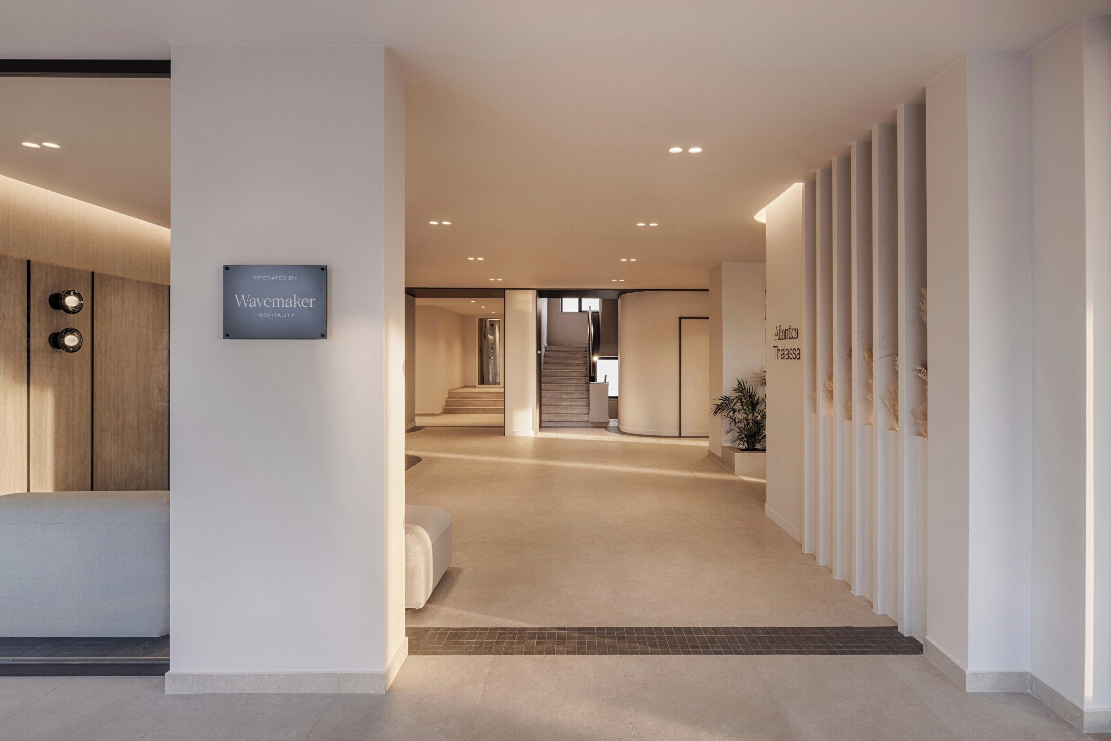 A modern hotel lobby with a neutral color palette, a reception desk, a staircase, and a sign indicating the hotel is operated by Wavemaker Hospitality.
