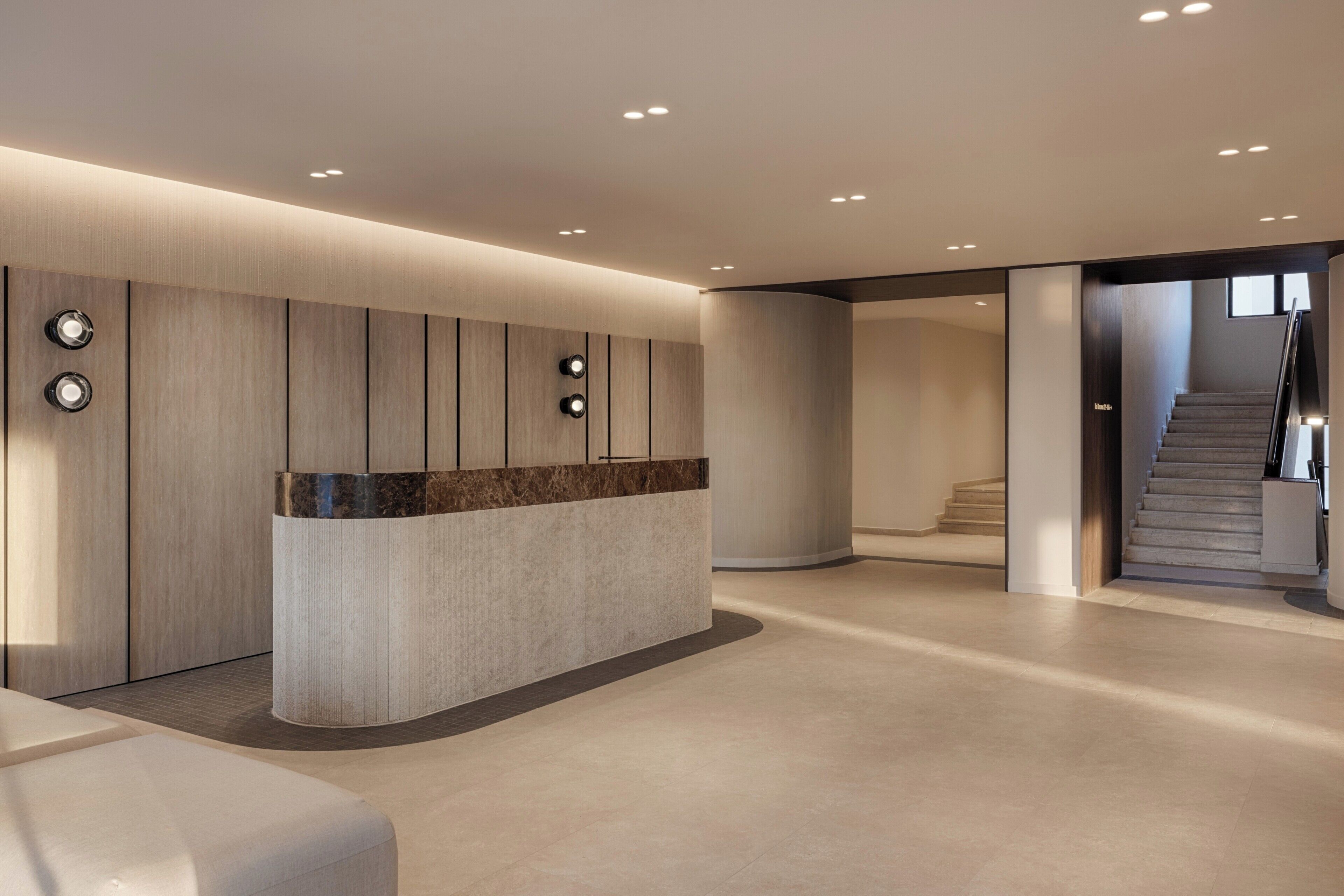 Un'area reception moderna con una scrivania a forma di arco, illuminazione a incasso e una scala che conduce a un piano superiore.