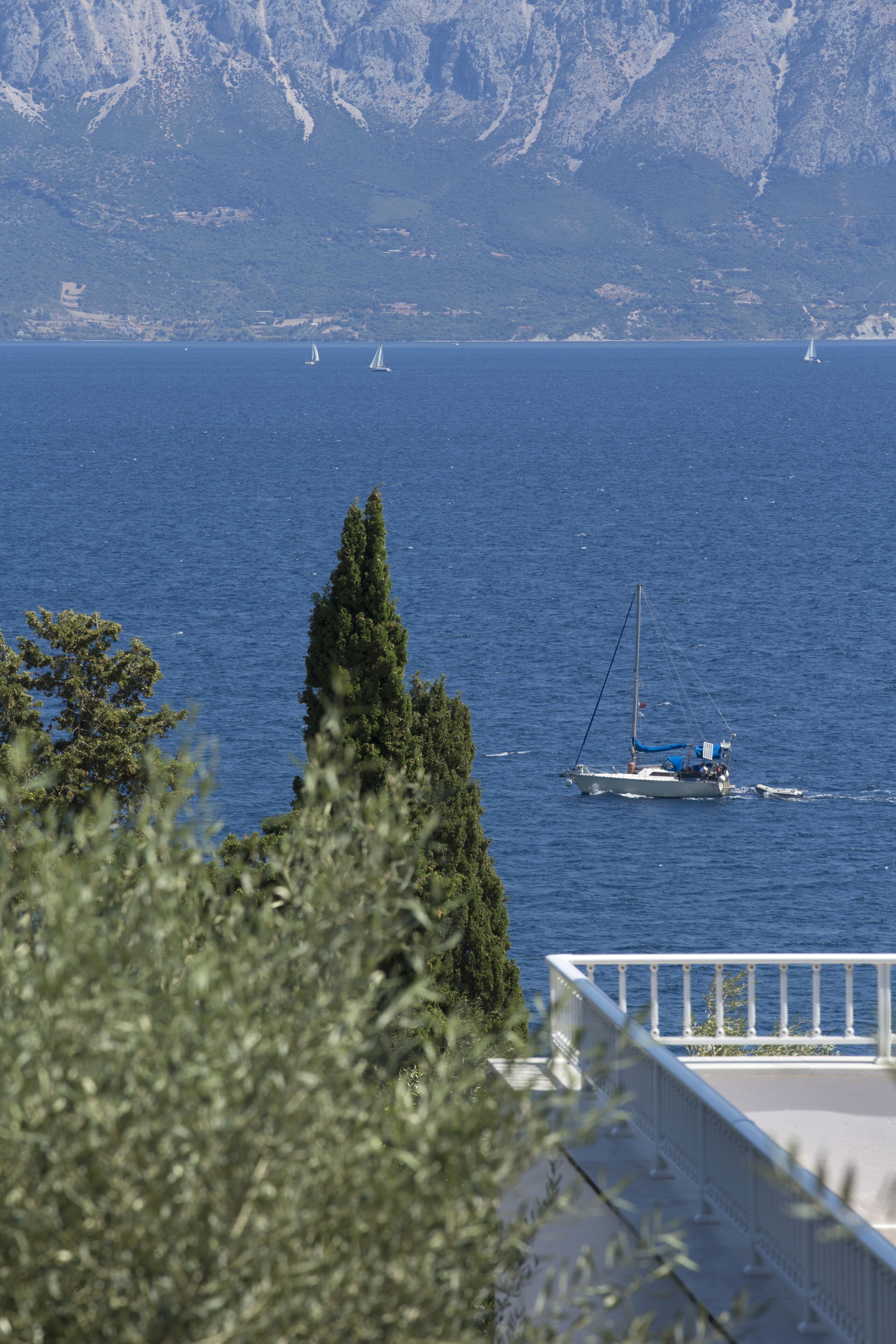 Une vue côtière avec un voilier sur l’eau, une montagne en arrière-plan et un bâtiment doté d’un balcon au premier plan.