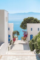 Un edificio blanco con puertas y ventanas azules, una escalera que lleva a una vista del mar, y un árbol con flores rosas.