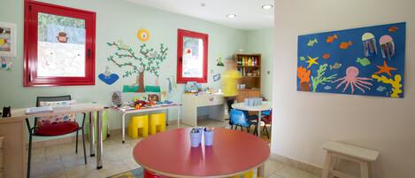 A brightly colored children's playroom with a red table, yellow and red chairs, and a blue wall mural.