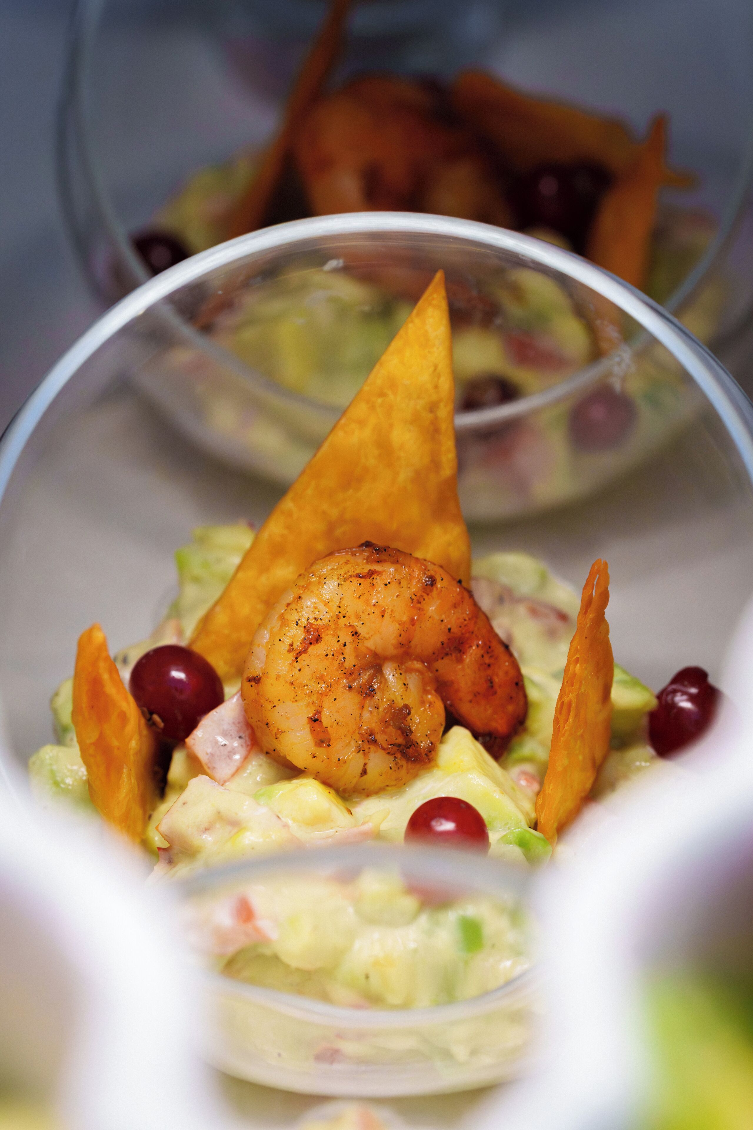 Un tazón de vidrio transparente con una ensalada de camarones, un totopo, un tomate cherry y una cucharada de salsa.