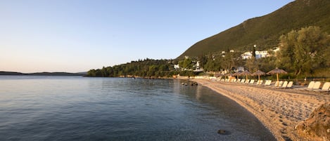 A pebble beach with clear water, a row of beach chairs, and buildings on a hillside.