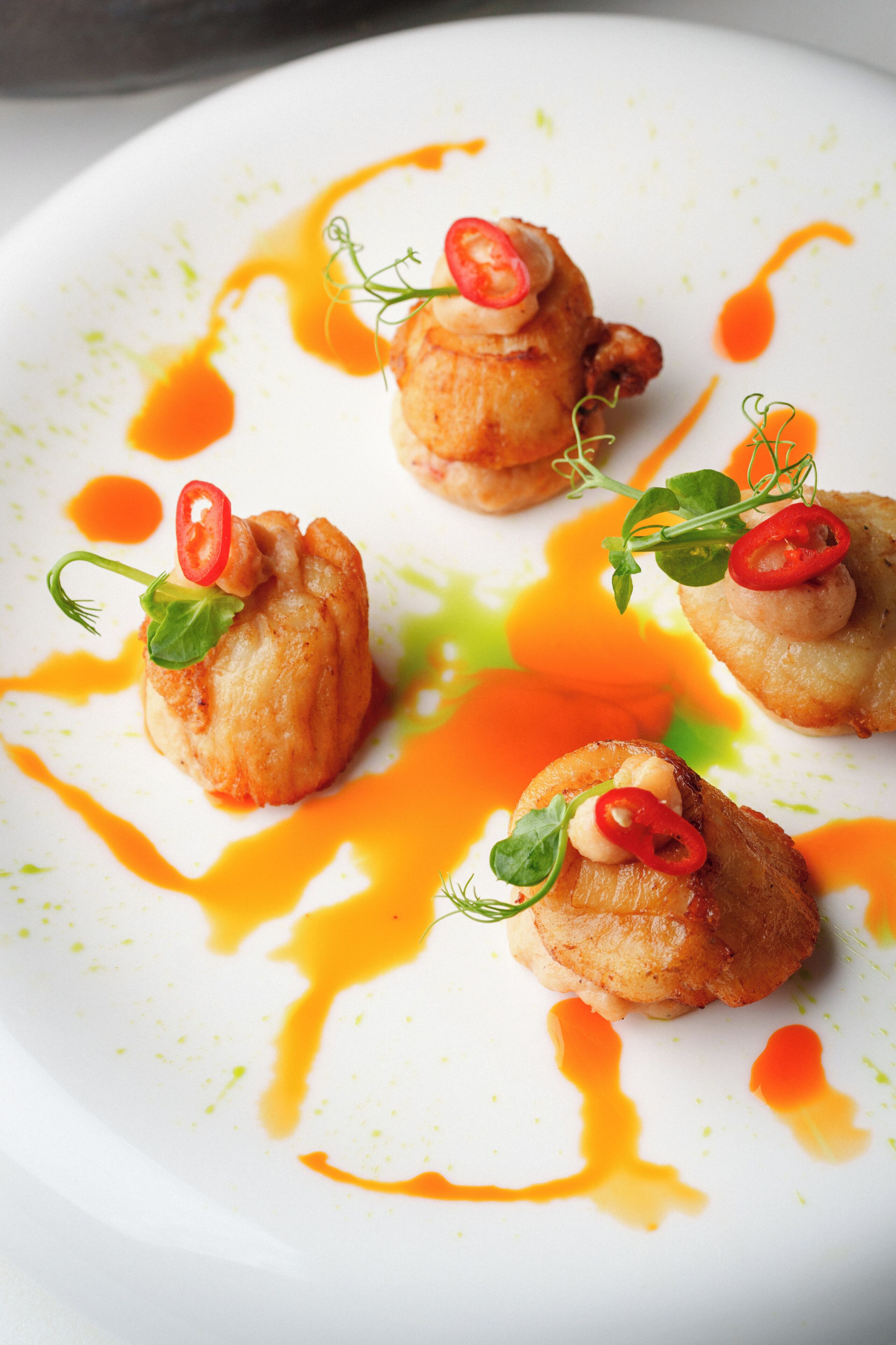 A white plate with four food items, each topped with a red chili slice and a green leaf, surrounded by orange sauce.