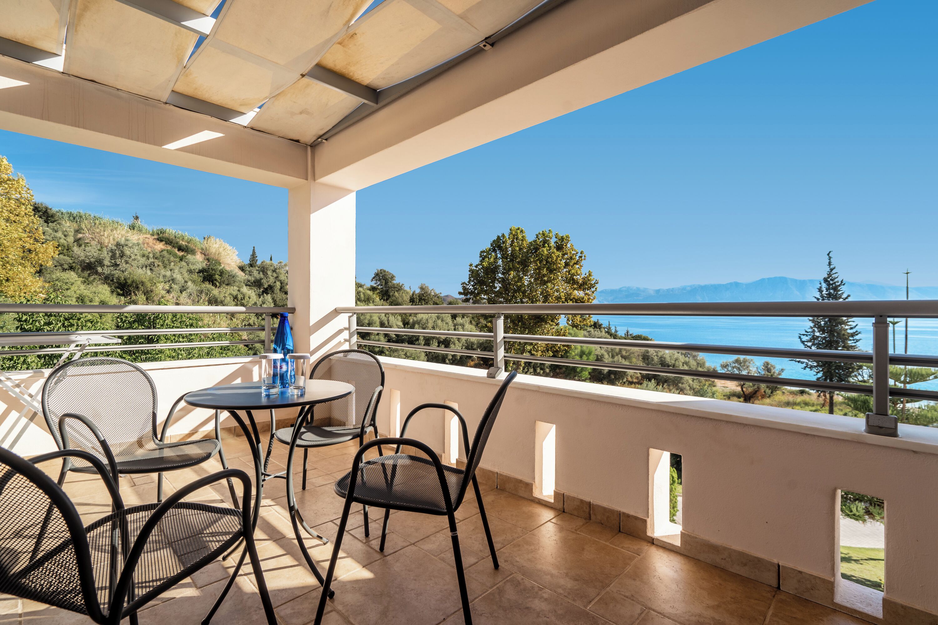 A balcony with outdoor furniture, a clear view of the sea, and a hilly landscape in the background.